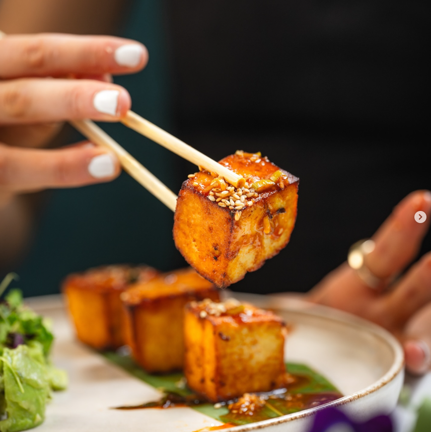 Chopsticks hold a sesame-topped, sauce-glazed tofu cube above a plate with more tofu and salad. Bright and appetizing scene.