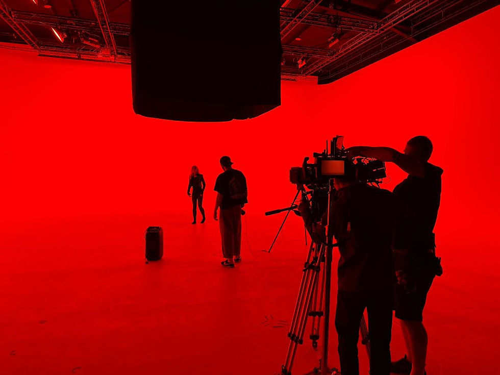 Three people in a red-lit studio. Two stand near a speaker, while one operates a camera. The atmosphere is intense and focused.