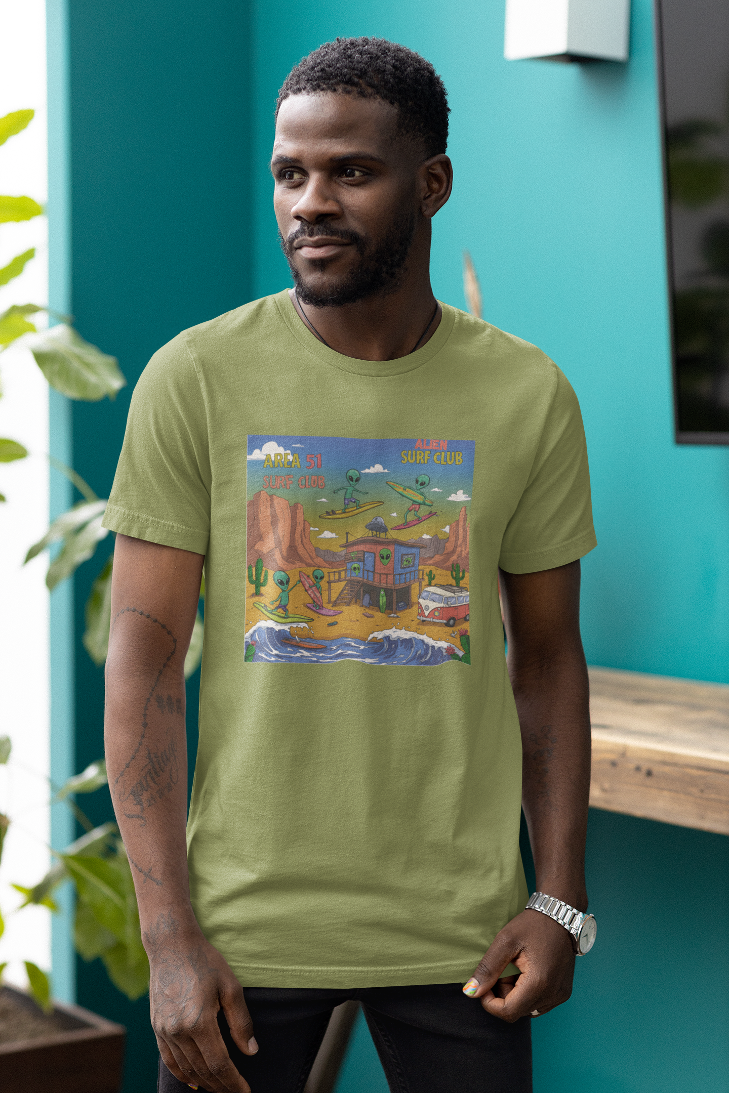 Man wearing a military green tee with a graphic of an alien surfing on a surfboard with Area 51 Surf Club