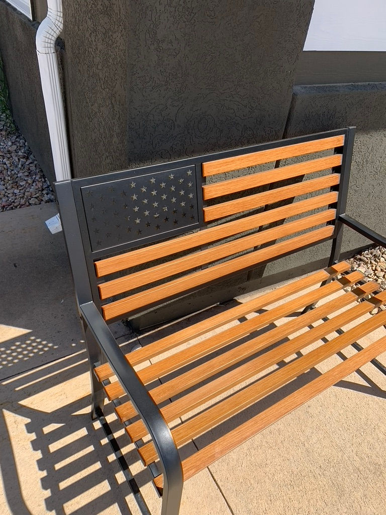 Wooden bench with black metal frame features a U.S. flag design on the backrest. It's placed on a concrete patio beside a building.