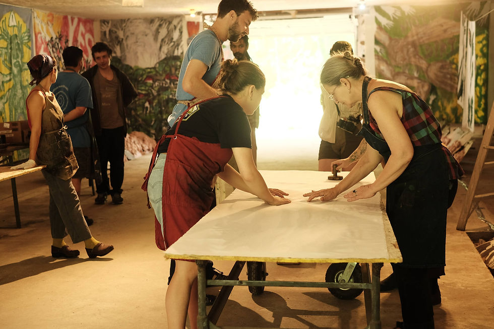 People in aprons work together on a large sheet at a table in a colorful art studio, with bright murals and a relaxed, collaborative vibe.