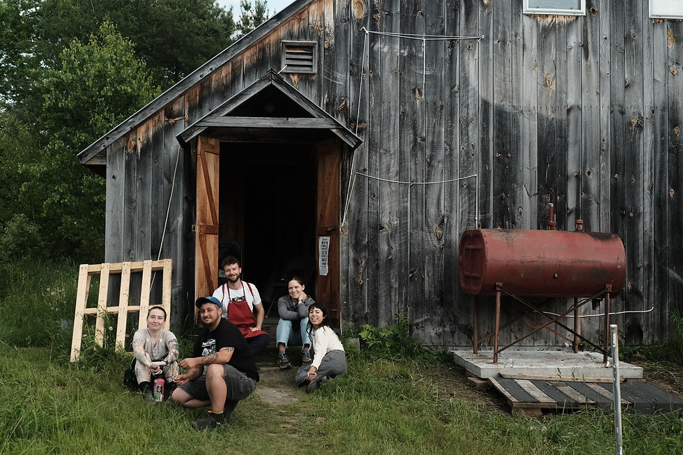 Six people smiling, sitting outside a rustic wooden building with a red tank. Grass and trees in the background. Relaxed, casual atmosphere.