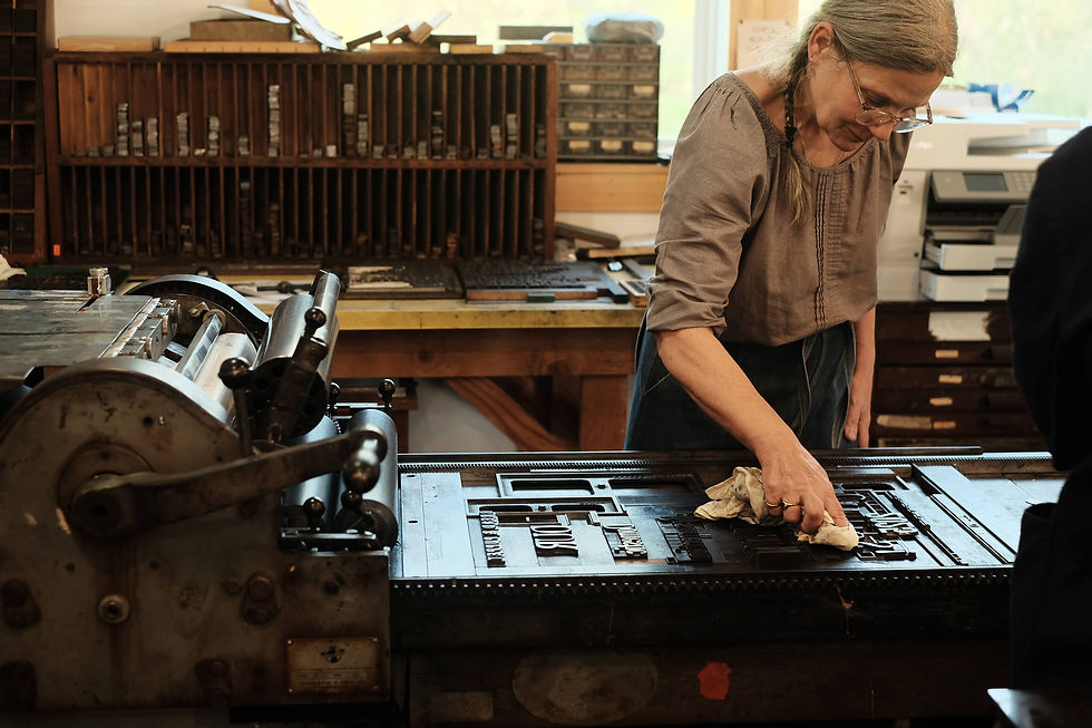 Woman cleaning a printing press in a workshop, surrounded by wood shelves and letter trays. The mood is focused and nostalgic.