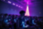 Audience seated with glowing blue devices in a dim room, illuminated by purple lighting. A large, lit tower structure in the background.