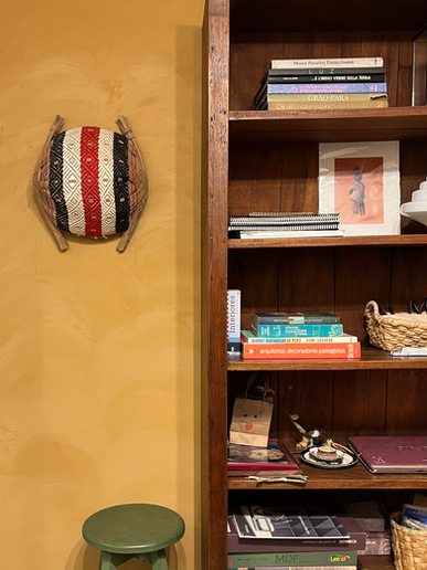 Composição de estante com livros e arte indígena, contrastando com a textura natural da parede em geotinta de barro e a presença de um cesto indígena artesanal
