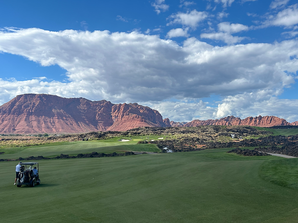 Golf course with a cart and players on a green field. Red rock mountains in the background beneath a partly cloudy blue sky.