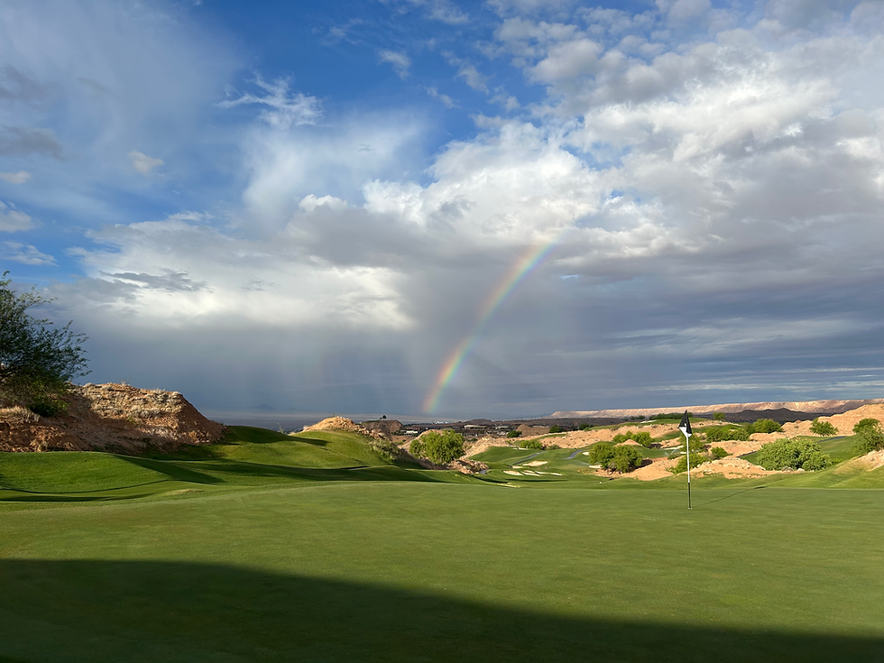 Golf course landscape with a rainbow in a cloudy sky, green fairway, and desert hills. A golf flag stands in the foreground. Peaceful mood.