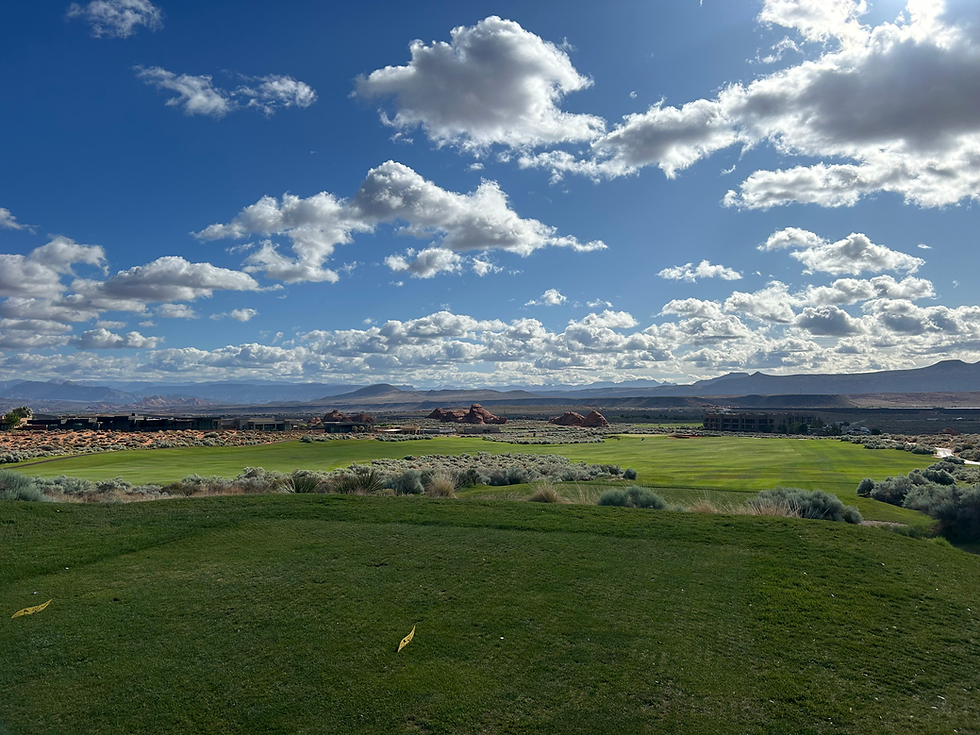 Green golf course under a bright blue sky with scattered clouds. Rolling hills and distant mountains create a serene backdrop.