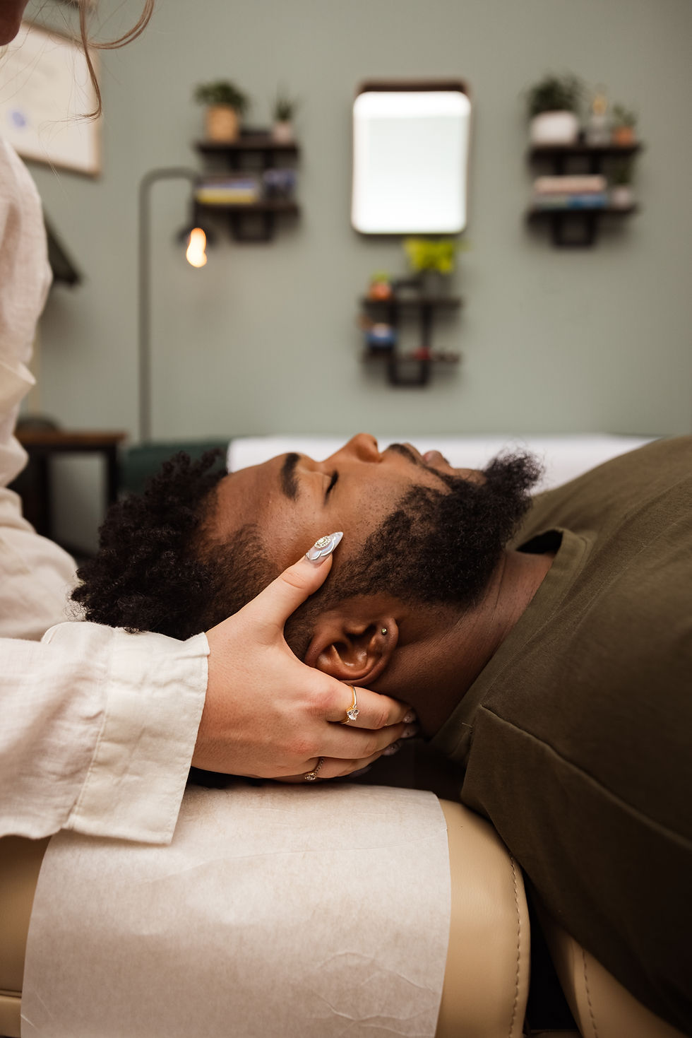 A person receives a massage and chiropractic adjustment lying on a table. Hands gently cradle the head. Room has green walls, shelves, and a mirror. Relaxing mood.
