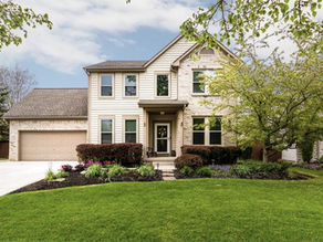 Two-story house with a beige facade, surrounded by green lawn and shrubs. Cloudy sky, trees in the background, calm suburban setting.