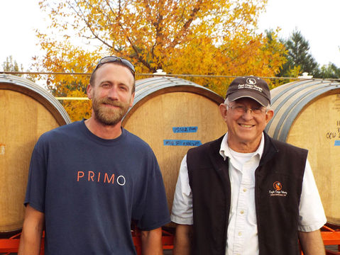 Two men smiling in front of wine barrels. One wears a "PRIMO" shirt, the other a cap and vest. Autumn trees in the background.