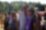 A smiling couple with sunglasses stands outdoors among a crowd. The woman wears a purple feather boa, and trees fill the sunny background.