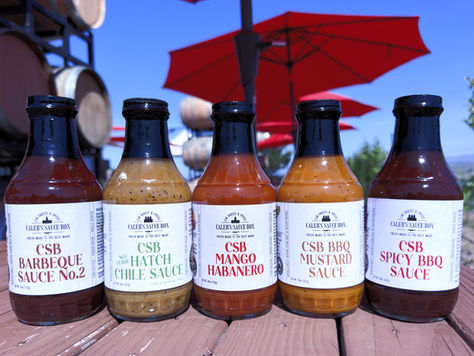 Five Caleb’s Sauce Box bottles of barbecue, hatch chile, mango habanero, mustard and spicy BBQ on a wooden table under red umbrellas.