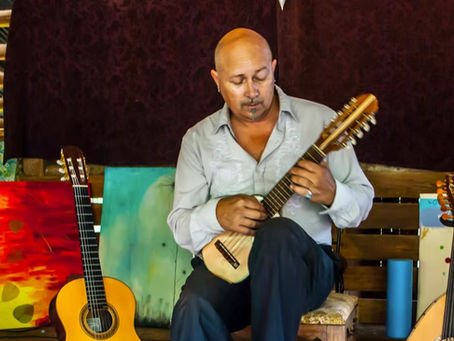 Andre Feriante seated playing a small string instrument among guitars in a room with vibrant artworks. Calm mood, colorful and rustic setting.
