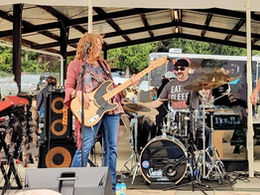 Outdoor band performs under a pavilion: singer at keyboard, bassist, drummer, and guitarist with blue guitar, upbeat live scene