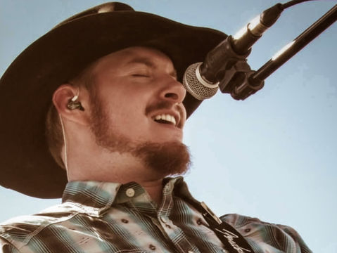 Country singer in a cowboy hat sings into a microphone while playing guitar against a clear blue sky.