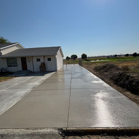 Freshly poured dark gray concrete slab drying in the bright sunlight with a white house in the distance. 