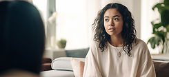 A girl with wavy black hair is sitting on a beige chair with pillows and talking to a person in the foreground.