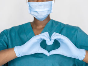 A medical professional in teal scrubs and a mask forms a heart shape with gloved hands, set against a plain white background. Well being screening at ReelCare Academy & Wellness..