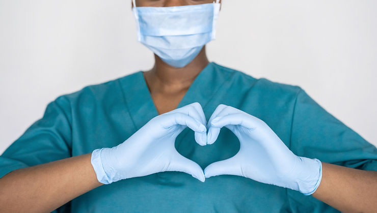 A medical professional in teal scrubs and a mask forms a heart shape with gloved hands, set against a plain white background. Well being screening at ReelCare Academy & Wellness.