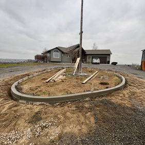 An dirt front yard with newly poured concrete curbing in the shape of an oval with a cloud covered sky overhead.