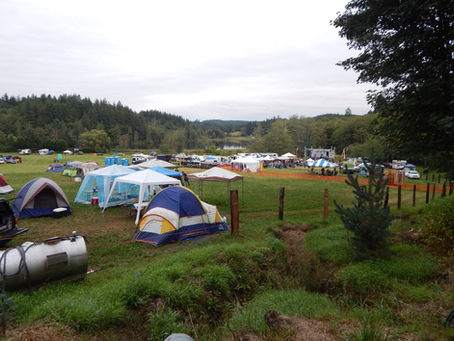 Campground festival with colorful tents, RVs, and a stage beside a lake, surrounded by forested hills under overcast skies.