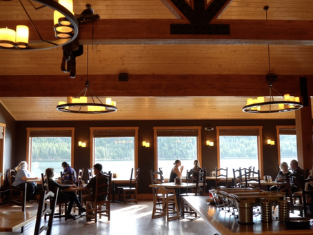 Half Moon Lake Lodge Dining Room. Cozy restaurant with wooden ceiling and chandeliers. People sitting at tables by large windows with scenic forest view outside. Warm ambiance. 