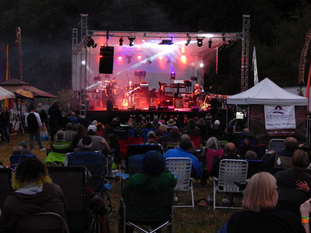 MoonFest's outdoor concert at dusk with band playing on a lit stage. Audience seated in lawn chairs under a partly cloudy sky.