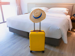 Yellow suitcase with straw hat in a bright hotel room. White bed, wooden floor, sheer curtains, and a cozy, inviting atmosphere.