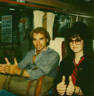Two people sit in a train, smiling and giving thumbs up. One wears glasses and a hat, the other a striped shirt. Text is visible in the background.