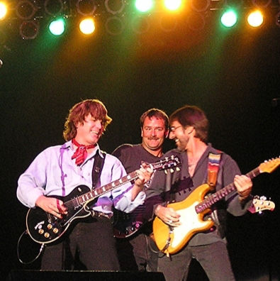 Three men play electric guitars on a dim stage under green, yellow, and orange lights, smiling and leaning together.