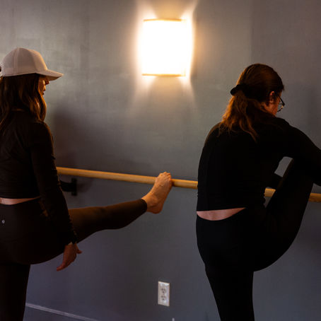 Three women stretch at a barre in a dimly lit studio. Background is gray with a glowing wall light.