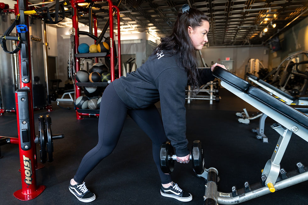 Woman lifting a dumbbell in a gym, wearing black athletic wear. She leans on a bench with focus. Background features equipment and weights.