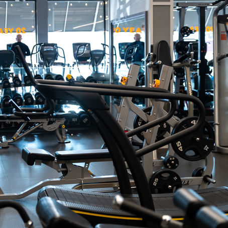A gym with various equipment, including treadmills and weights. Bright lighting through windows; text "20 YEARS" seen on glass.
