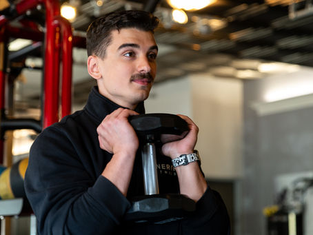 Man in gym holds a dumbbell close to his chest, wearing a black jacket with white text and a patterned watch. Bright, modern background.