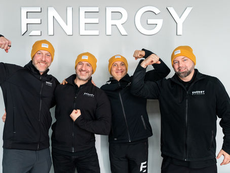 Four smiling men in black jackets and yellow beanies flex muscles under the bold text "ENERGY" on a gray wall.