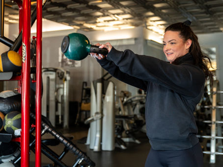 Woman in a black top lifts a green kettlebell in a gym. Background features exercise equipment and workout balls. Focused expression.
