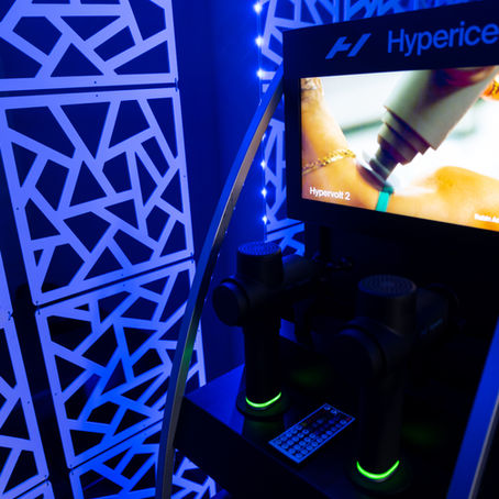 Hyperice display with massage devices, screen shows massage tool in use. Blue geometric background, vibrant lighting creates energetic mood.