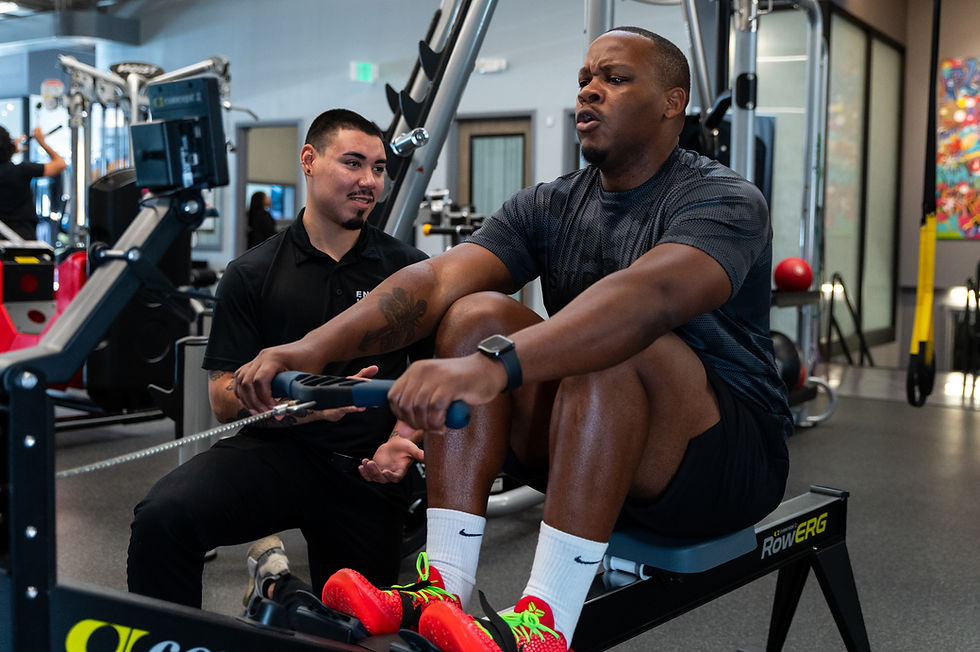 Man exercising on a rowing machine, trainer kneeling beside him. Gym setting, bright colors from machines and art. Focused and determined mood.