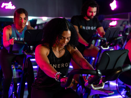 People in a dimly lit gym engage in a spin class. Neon signs glow in the background. The word "ENERGY" is visible on their shirts.