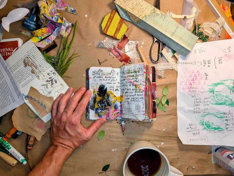 a hand holds a Pocket Atelier open on a messy desk