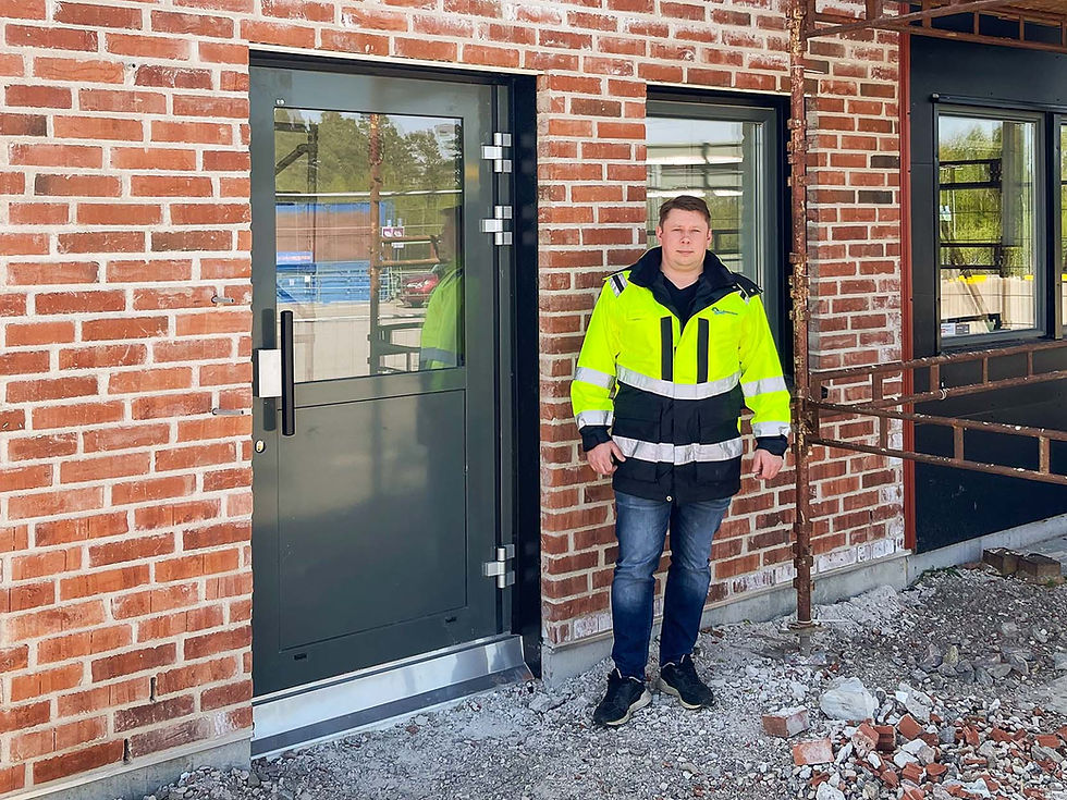 A man in a high-visibility jacket stands in front of a building under construction. He is outside a glass door framed by a brick wall with visible sections of broken bricks at his feet. The building has a modern design with a combination of brick and black metal frames. The man looks directly at the camera, suggesting he might be involved in the construction project.