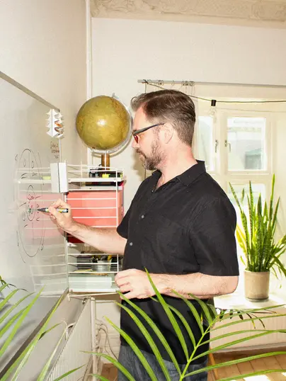 En man ritar på en whiteboard, klädd i svart skjorta och står i ett rum med växter och en stor jordglob. Han verkar engagerad i att skissa upp en diagram, med ett tankfullt uttryck, vilket antyder en brainstormingsession eller planering.