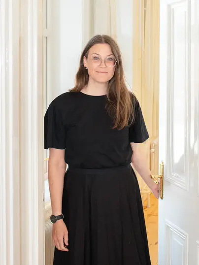 En kvinna klädd i svart tröja och kjol står självsäkert i en dörröppning. Hon har långt, rakt hår, runda glasögon och ler varmt mot kameran. Rummet i bakgrunden är mjukt upplyst, vilket skapar en känsla av värme och öppenhet.