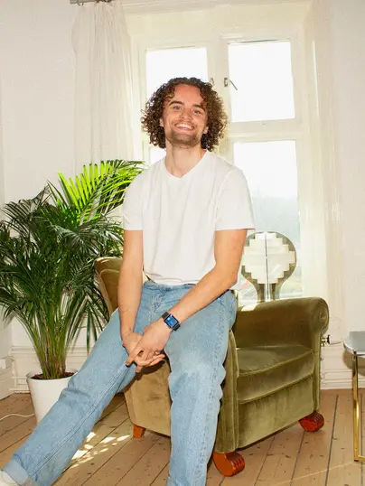 En leende ung man med lockigt hår sitter avslappnat på en grön fåtölj i ett ljust rum. Han är klädd i en vit t-shirt och ljusa jeans. Bakom honom finns en stor grön växt och ett fönster som släpper in dagsljus. Den avslappnade hållningen och det varma leendet ger en känsla av trivsel och harmoni i den luftiga miljön.