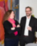 En kvinna och en man står leende i ett samtal. Kvinnan har långt brunt hår och bär en svart tröja samt en rosa sjal. Mannen har glasögon, kort skägg och är klädd i en mörk kavaj över en vit skjorta. Han håller en vit kopp i handen. I bakgrunden syns en färgglad textilvägg.