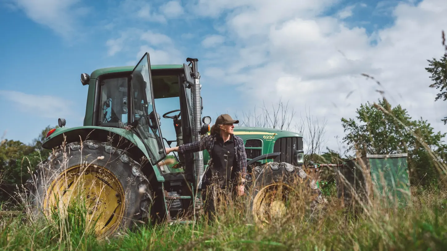 En kvinna klädd i arbetskläder står vid en grön traktor på en gräsbevuxen åker under en blå himmel.