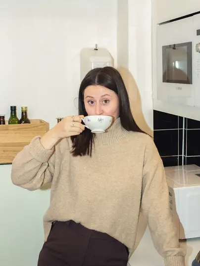 En kvinna i en beige tröja dricker ur en blommig tekopp medan hon lutar sig avslappnat mot en köksbänk. Bakom henne syns olika flaskor med kryddor och en mikrovågsugn, vilket ger en hemtrevlig känsla.