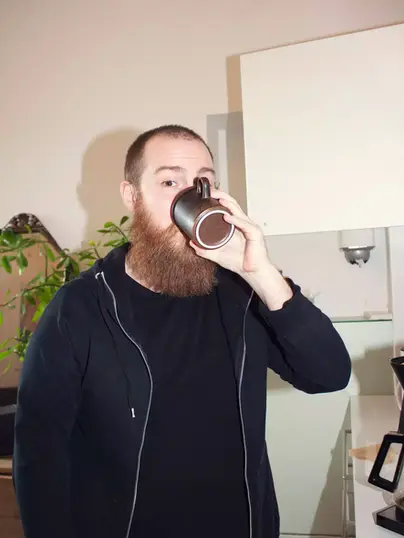 En person med mörk skägg och svart hoodie dricker ur en mörk kaffemugg i ett kök. I bakgrunden syns en kaffebryggare, ett skåp och en grön växt.