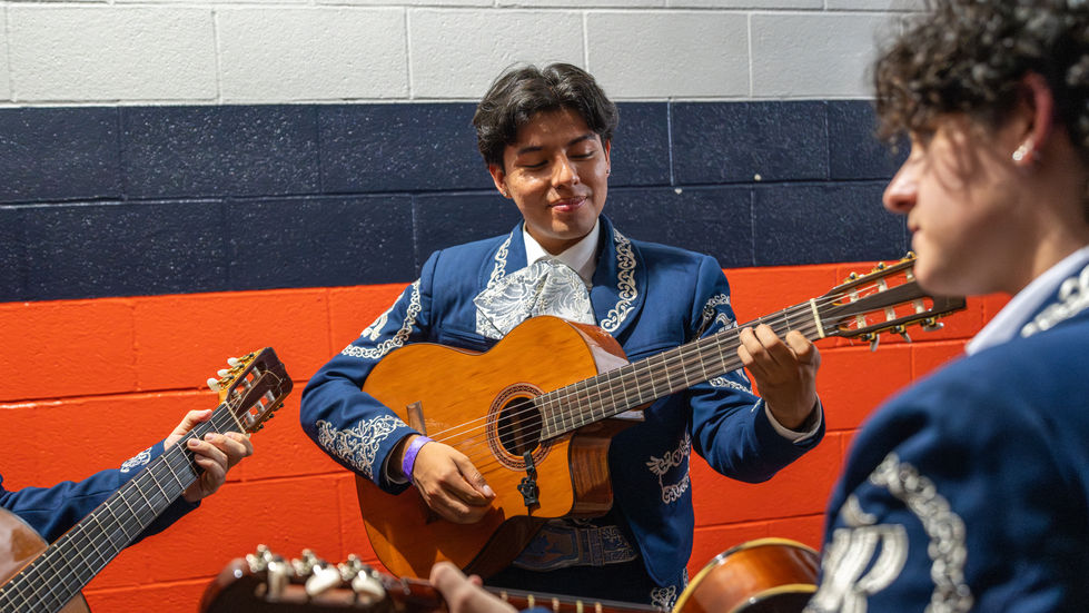 A guitar player plays his guitar while two other mariachi members are in the foreground facing him, inside the tunnels of Daikin Park waiting to play